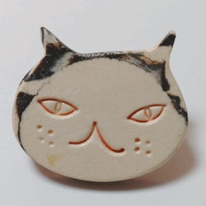 以下が含まれることがあります： 耳と額に黒いペイントが施された、オフホワイトの猫型セラミックブローチ。アーモンド型の目、鼻、ひげの斑点などの顔のパーツは表面に彫り込まれており、オレンジ色のディテールで強調されています。