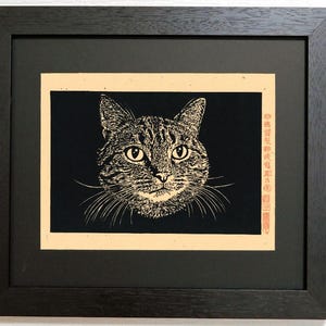 以下が含まれることがあります： 猫の顔を描いた黒とクリーム色のコントラストが強いイラストの額入りプリント。猫には詳細なひげと縞模様がある。猫の右側には、赤いインクで縦書きの日本語のテキストと、赤い四角い印章が押されている。