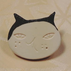 以下が含まれることがあります： クリーム色の顔と黒い耳を持つ猫の頭の形をした陶器製のブローチ。猫には彫り込まれたアーモンド型の目、小さな曲がった口、そして両頬に4つの小さな点が2組あり、独特の表情を作り出しています。