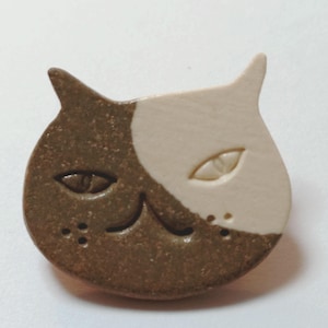 Op de afbeelding: Een keramische broche in de vorm van een kat met een tweekleurig ontwerp, een gespikkelde bruine afwerking aan de linkerkant en een gladde crèmekleurige afwerking aan de rechterkant. Het kattengezicht heeft ingesneden amandelvormige ogen en een mond.