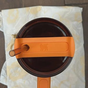 Pu&ograve; includere: Vista dall'alto di un bicchiere marrone con manico e coperchio arancione. Il coperchio ha una cannuccia e una fascia arancione con un logo. Il bicchiere poggia su un panno bianco.
