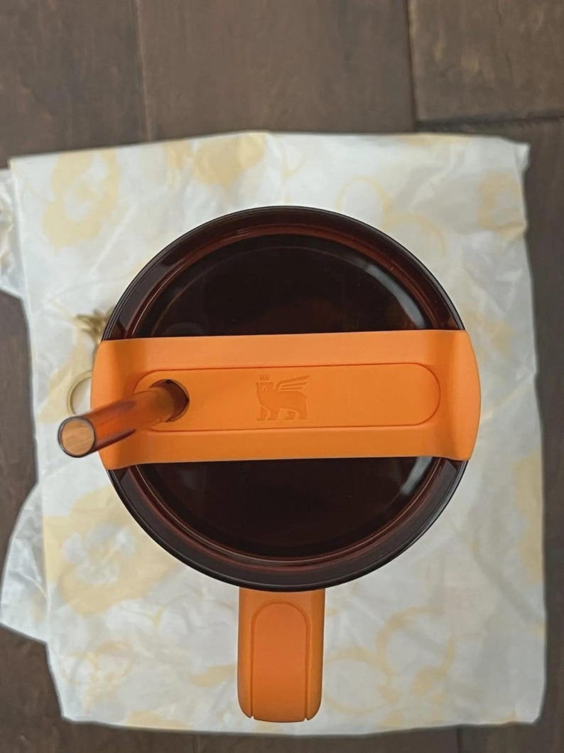 Pu&ograve; includere: Vista dall'alto di un bicchiere marrone con manico e coperchio arancione. Il coperchio ha una cannuccia e una fascia arancione con un logo. Il bicchiere poggia su un panno bianco.