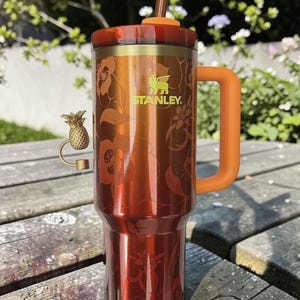 Pu&ograve; includere: Un grande bicchiere termico con finitura color rame e design floreale. Ha un manico e coperchio arancioni, una cannuccia marrone e un ciondolo a forma di ananas dorato. Il logo Stanley &egrave; visibile.