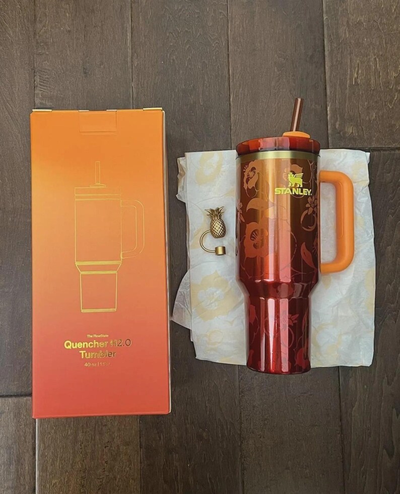 Pu&ograve; includere: Un bicchiere Stanley Quencher 2.0 rosso e oro con manico e cannuccia, accanto alla sua scatola arancione e oro. La scatola dice "Quencher 112.0 Tumbler 1183 ml". &Egrave; presente anche un ciondolo a forma di ananas dorato.
