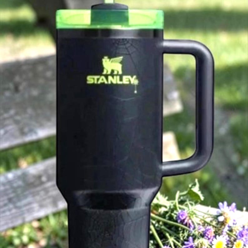 K&ouml;nnte beinhalten: Schwarzer Stanley-Reisebecher mit gr&uuml;nem Deckel und Griff. Das Stanley-Logo ist gelb. Der Becher steht auf einer Holzbank mit Blumen im Hintergrund.