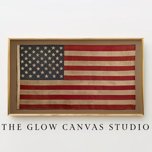 May include: Framed American flag with red and white stripes and a blue field of white stars. The flag is displayed in a gold-colored frame. The text "THE GLOW CANVAS STUDIO" is printed below the frame.