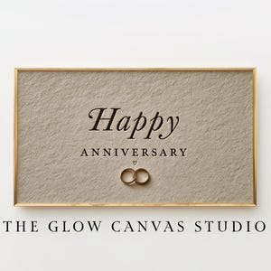 May include: A rectangular framed print with a textured beige background featuring the dark brown text Happy ANNIVERSARY and a small heart symbol above two interlocking gold rings. The frame is thin and metallic gold in color.