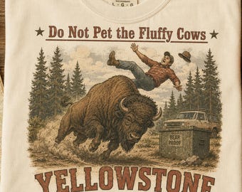 May include: A cream-colored t-shirt featuring a graphic of a person being tossed into the air by a large bison. The text reads Do Not Pet the Fluffy Cows and Yellowstone National Park. A bear-proof container and a truck are visible in the background.