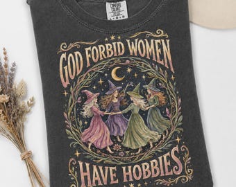 May include: A charcoal gray crew neck t-shirt featuring a graphic print of four figures in witch hats and long dresses dancing in a circle. The text reads GOD FORBID WOMEN HAVE HOBBIES. The design includes a crescent moon, stars, and foliage.
