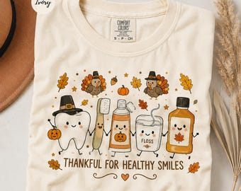May include: An ivory t-shirt featuring a graphic of a smiling tooth, toothbrush, toothpaste, floss, and mouthwash, all wearing pilgrim hats or decorated with autumn leaves and pumpkins. The text reads, Thankful for Healthy Smiles, with turkeys.