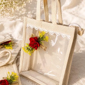 May include: A rectangular tote bag made of light beige burlap with two matching handles and a clear plastic front panel. The bag features a white lace trim along the top edge and a decorative cluster of red fabric flowers and yellow sprigs.