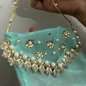 Può includere: Un accessorio in tessuto trasparente color turchese chiaro con bordo metallico dorato a forma di foglia, decorato con perle artificiali bianche e piccole paillettes dorate a motivi floreali, sospeso da un sottile cinturino dorato con perle.