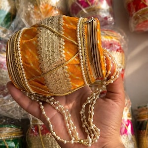 May include: A cylindrical yellow fabric pouch with a white tie-dye pattern, featuring a gold metallic lace trim, a zipper closure, and a long beaded strap made of small white pearls. The pouch is decorated with vertical gold cord accents.