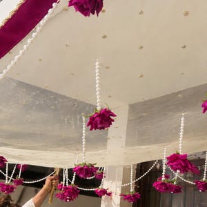 Può includere: Un baldacchino in tessuto bianco con un bordo magenta intenso e fili di perle artificiali bianche pendenti. Ogni filo di perle è decorato con una rosa artificiale color magenta vibrante all'estremità, creando un ornamento decorativo.