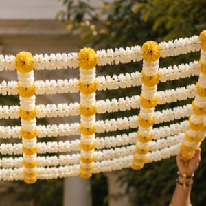 May include: A decorative garland constructed from a grid of white and yellow flowers. The design features vertical and horizontal strands of small white blossoms intersected by larger, round, vibrant yellow marigold-like flower clusters.