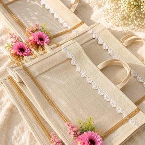 May include: A set of rectangular beige jute tote bags featuring a clear plastic window on the front, white lace trim along the edges, and a decorative cluster of pink artificial flowers with green foliage attached to the bottom corner.