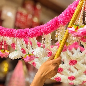 May include: A decorative floral garland featuring vibrant pink and white artificial flowers arranged in dense clusters. The garland is accented with strings of small white faux pearls and is attached to a gold-colored rod for display purposes.