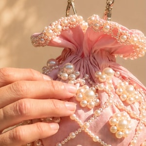 May include: A light pink drawstring pouch bag featuring an intricate lattice pattern of white faux pearls. The bag is adorned with floral-shaped pearl clusters and a long fringe of dangling pearl beads at the base, finished with a pearl bead handle.