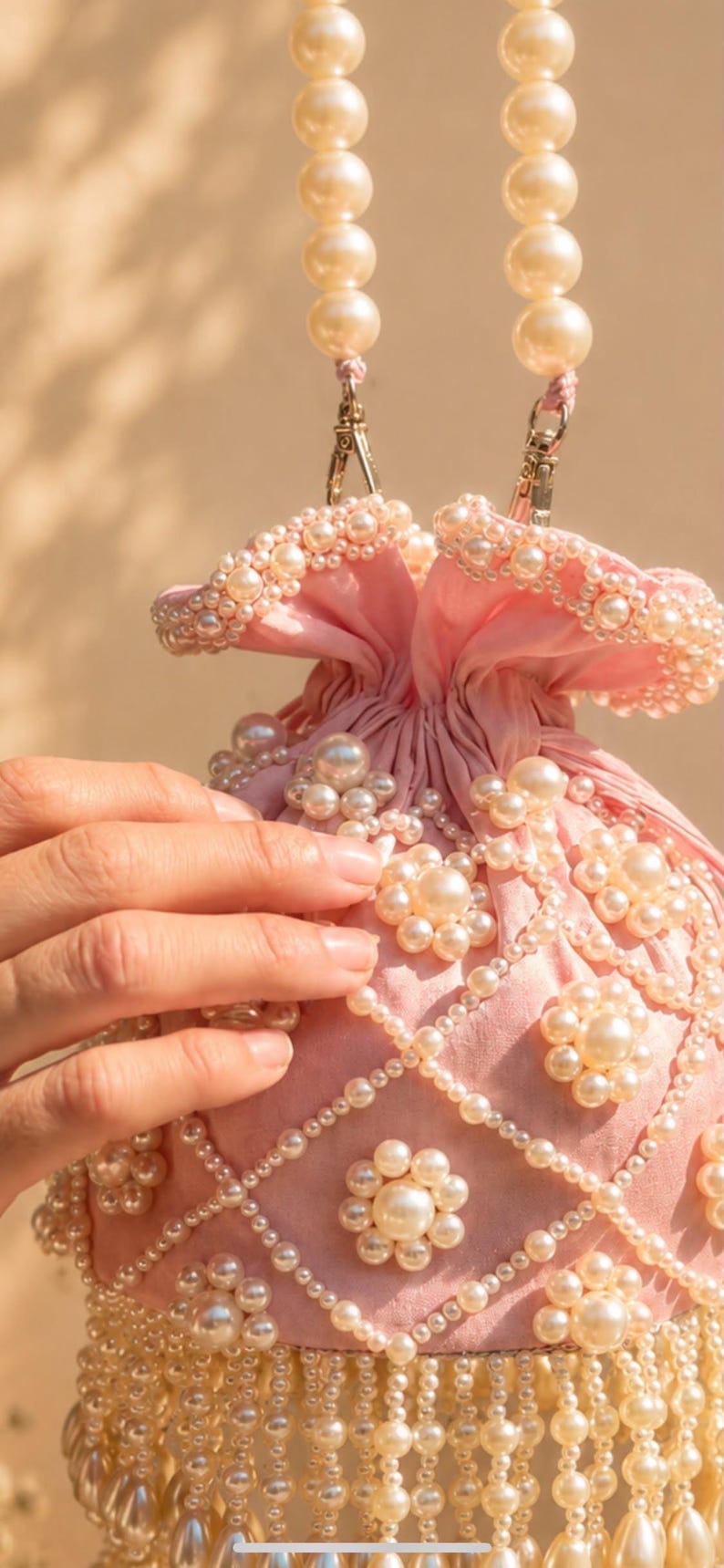 May include: A light pink drawstring pouch bag featuring an intricate lattice pattern of white faux pearls. The bag is adorned with floral-shaped pearl clusters and a long fringe of dangling pearl beads at the base, finished with a pearl bead handle.