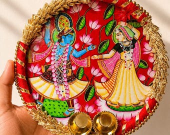 Può includere: Un vassoio decorativo circolare con uno sfondo rosso vivace e un'illustrazione dipinta di due figure in abiti tradizionali tra fiori di loto rosa. Il bordo è decorato con frange metalliche dorate e due piccole ciotole dorate.