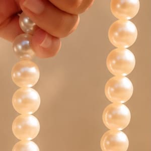 May include: A strand of round, cream-colored pearls strung together. The pearls have a smooth, lustrous surface and are uniform in size. At the bottom of the strand, a small metal clasp is attached with a light pink thread, securing the pearls.