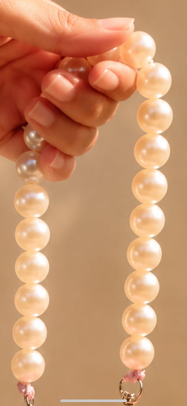 May include: A strand of round, cream-colored pearls strung together. The pearls have a smooth, lustrous surface and are uniform in size. At the bottom of the strand, a small metal clasp is attached with a light pink thread, securing the pearls.