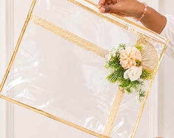 May include: A rectangular clear plastic storage bag featuring gold-toned trim along the edges and a zipper closure. The front of the bag is decorated with an arrangement of artificial peach and white flowers, green foliage, and a gold fan-shaped accent.