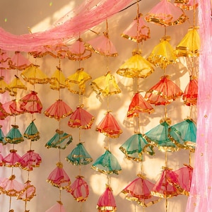 May include: A decorative hanging garland featuring multiple vertical strands of small, cone-shaped fabric ornaments in pink, yellow, red, and teal. Each ornament is trimmed with metallic gold ribbon and suspended from a sheer pink fabric drape.