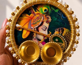 Può includere: Un piatto decorativo circolare con una colorata illustrazione di Krishna con una piuma di pavone, decorato con un bordo avvolto in filo dorato, perle bianche e perline dorate. Due piccole ciotole dorate sono fissate sulla superficie frontale.