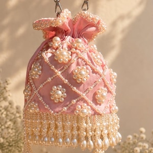 May include: A light pink drawstring pouch bag featuring a lattice pattern of white faux pearls. The bag is adorned with floral-shaped pearl clusters and a dense fringe of dangling teardrop and round pearls at the base, with a pearl handle.