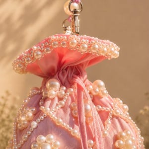 May include: A light pink drawstring pouch bag featuring a gathered top and a handle made of large white faux pearls. The exterior is decorated with intricate patterns of white faux pearls arranged in floral shapes and connected by beaded strands.