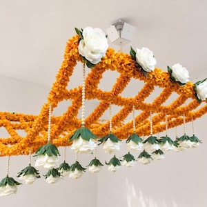 Puede incluir: Una decoración de techo colgante construida con una red de flores de caléndula naranjas. Flores de rosa blancas están unidas al marco superior y suspendidas de la estructura mediante hilos de pequeñas cuentas blancas, creando un arreglo floral.