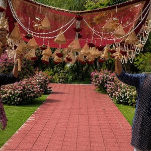 May include: A rectangular deep red fabric canopy featuring intricate gold embroidery along the edges. The canopy is adorned with multiple hanging beige cone-shaped tassels, strings of white faux pearls, and small dark red floral accents.