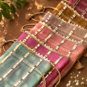 Könnte beinhalten: Eine Sammlung rechteckiger Beutel aus transparentem Stoff in verschiedenen Farben wie Türkis, Pink und Gold. Jeder Beutel ist mit vertikalen und horizontalen goldfarbenen Metallbändern mit gezacktem Rand sowie einem Kordelzug mit kleinen Quasten versehen.