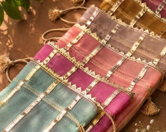 May include: A collection of rectangular sheer fabric pouches in various colors including teal, pink, and gold. Each pouch features vertical and horizontal gold metallic ribbon trim with a serrated edge and a drawstring closure with small gold tassels.