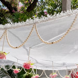 May include: A white parasol featuring a delicate lace trim along the edge, adorned with draped strings of cream-colored faux pearls and small, artificial pink rose blossoms hanging from the canopy in a decorative, symmetrical arrangement.