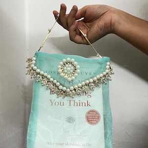 Op de afbeelding: Een licht turquoise doorzichtige stoffen boekomslag met een decoratieve rand van witte imitatieparels en goudkleurige metalen blaadjes. De hoes heeft een bijpassend handvat met parels en bevat het boek "Everything You Think".