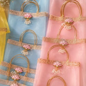 May include: A collection of light blue and soft pink organza gift bags featuring gold metallic handles, decorative gold lace trim, and a central pink lotus flower embellishment with dangling white faux pearl beads attached to the front.