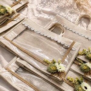 May include: A collection of rectangular beige tote bags made of woven fabric, each featuring a clear plastic window on the front, a white lace trim along the top edge, gold ribbon accents, and a cluster of artificial green and white flowers.