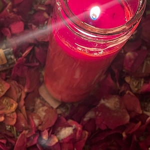 Puede incluir: Una vela roja encendida en un tarro de cristal transparente, rodeada de p&eacute;talos de rosa rojos secos. La llama de la vela brilla intensamente, creando una luz c&aacute;lida. La escena evoca un ambiente m&iacute;stico.