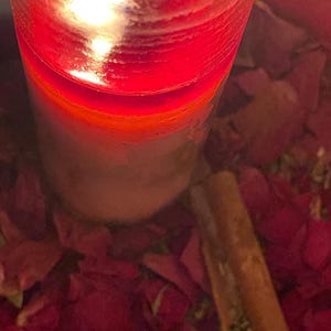 Puede incluir: Una vela roja encendida en un recipiente de vidrio transparente. La vela est&aacute; rodeada de p&eacute;talos de flores rojas y una ramita de canela. La llama es brillante y la cera tiene un degradado de rojo y blanco.