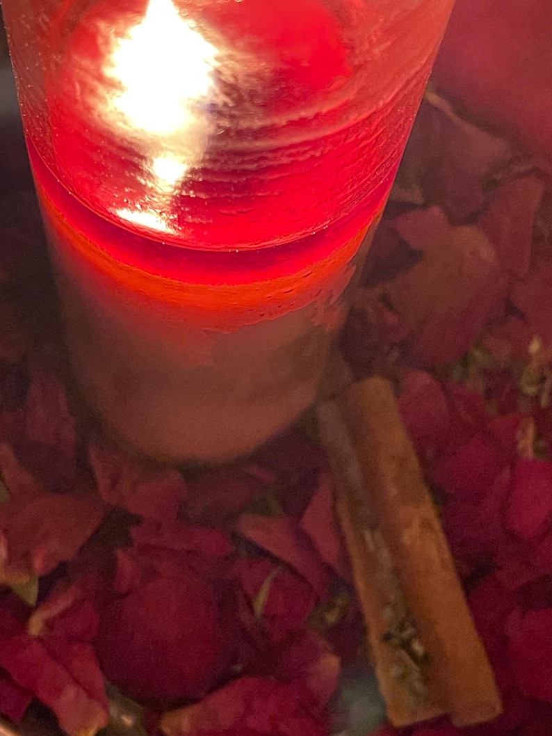 Puede incluir: Una vela roja encendida en un recipiente de vidrio transparente. La vela est&aacute; rodeada de p&eacute;talos de flores rojas y una ramita de canela. La llama es brillante y la cera tiene un degradado de rojo y blanco.