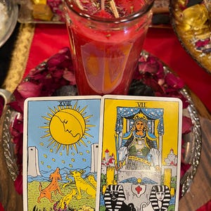 Puede incluir: Una vela roja en un tarro de cristal, con cartas del tarot que muestran "La Luna" y "El Carro". Las cartas est&aacute;n sobre una bandeja plateada con p&eacute;talos de rosa secos. La vela tiene un dise&ntilde;o en forma de escudo.