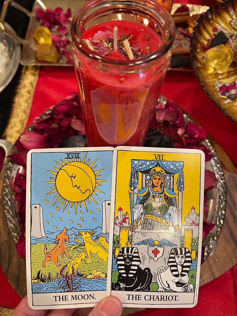 Puede incluir: Una vela roja en un tarro de cristal, con cartas del tarot que muestran "La Luna" y "El Carro". Las cartas est&aacute;n sobre una bandeja plateada con p&eacute;talos de rosa secos. La vela tiene un dise&ntilde;o en forma de escudo.