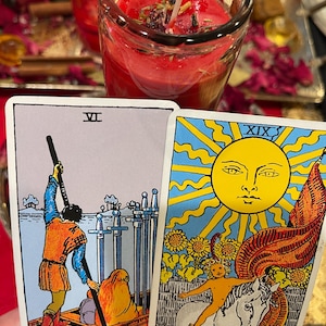 Puede incluir: Dos cartas de tarot, una que representa una figura en un barco con espadas, la otra que muestra un sol con una figura en un caballo. Una vela roja en un frasco de vidrio est&aacute; en el fondo. La carta dice "THE SUN."