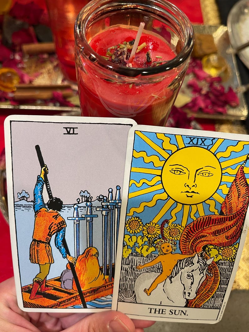 Puede incluir: Dos cartas de tarot, una que representa una figura en un barco con espadas, la otra que muestra un sol con una figura en un caballo. Una vela roja en un frasco de vidrio est&aacute; en el fondo. La carta dice "THE SUN."
