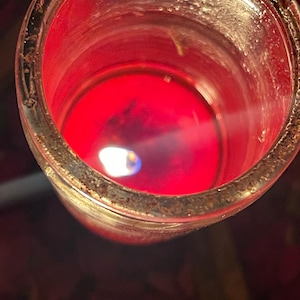 Puede incluir: Una vela roja encendida en un tarro de cristal transparente. La peque&ntilde;a llama tiene un n&uacute;cleo azul. El borde del tarro tiene un aspecto texturizado de color marr&oacute;n dorado. La cera de la vela es de un rojo intenso y vibrante.
