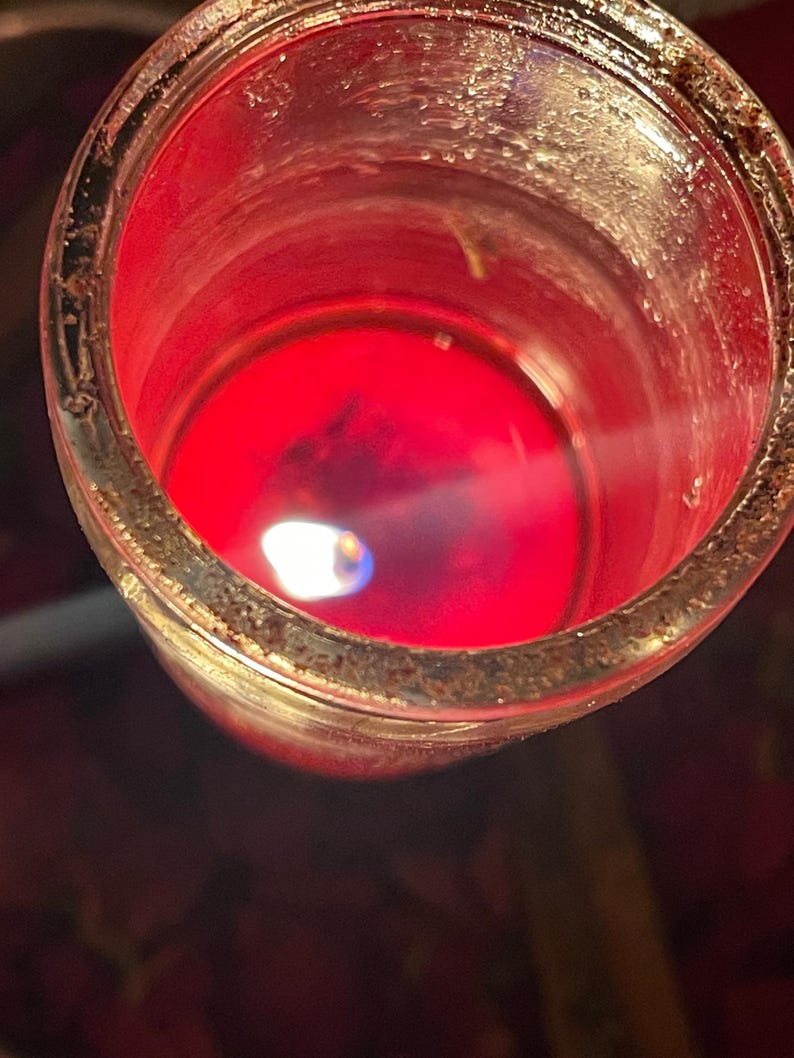 Puede incluir: Una vela roja encendida en un tarro de cristal transparente. La peque&ntilde;a llama tiene un n&uacute;cleo azul. El borde del tarro tiene un aspecto texturizado de color marr&oacute;n dorado. La cera de la vela es de un rojo intenso y vibrante.