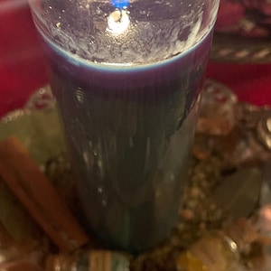Puede incluir: Una vela azul oscuro encendida en un vaso transparente. La vela se encuentra sobre una bandeja decorativa plateada con palitos de canela, piedras peque&ntilde;as y una botella peque&ntilde;a. La llama de la vela es brillante y la cera tiene una apariencia texturizada.