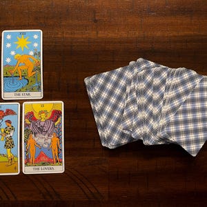 Puede incluir: Una tirada de cartas del tarot con tres cartas boca arriba y una baraja de cartas. Las cartas muestran ilustraciones coloridas con t&iacute;tulos como "The Star" y "The Lovers". La baraja tiene un patr&oacute;n de cuadros azules y blancos en el reverso.